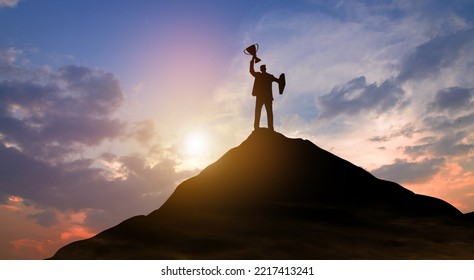 Silhouette Victory Businessman With Trophy Climbed To The Top Of Mountain Over The Sky And Sun Light Background. Success, Achieving Goal, Leadership, Career, Winner, And Business Achievement Concept.