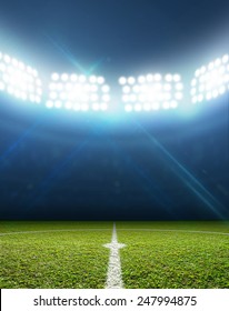 A Soccer Stadium With A Marked Green Grass Pitch With A Soccer Ball On The Center Mark At Night Under Illuminated Floodlights