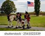 Soldiers in traditional War of 1812 military uniforms playing fife and drum, framed by the iconic American flag waving under a bright, sunny sky.