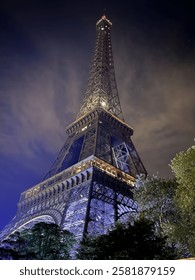 A stunning nighttime view of the Eiffel Tower, illuminated against a dark sky with clouds. The warm and cool lighting highlights the intricate iron structure, creating a captivating cityscape shot.