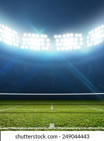 A Tennis Court In An Arena With A Marked Green Lawn Surface At Night Under Illuminated Floodlights