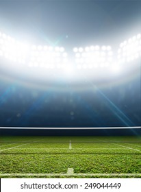 A Tennis Court In An Arena With A Marked Green Lawn Surface At Night Under Illuminated Floodlights