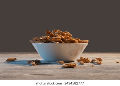 The textured surface of a rustic wooden table highlights a white bowl overflowing with fresh almonds, surrounded by a scattering of nuts on a subdued grey backdrop. - Powered by Shutterstock