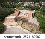 This aerial photograph captures an old stone fortification surrounded by lush greenery and a small town in the background. The fort features thick stone walls, red-tiled roofs, and a central courtyard