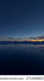 A tranquil 3d Rendering of a star-filled sky over jagged mountains and a glassy lake at dusk.
