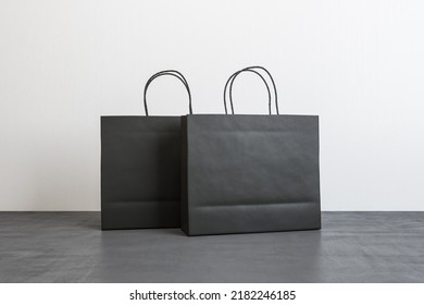 Two Blank Black Paper Shopping Bags With Place For Your Logo Or Text On Concrete Floor On White Wall Background. 3D Rendering, Mockup