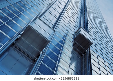 Two elevators are moving up on the side of a modern skyscraper. Blue sky reflects in the windows. The building is located in a bustling urban city, showcasing modern architecture and design - Powered by Shutterstock