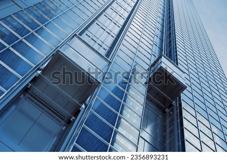 Two elevators are moving up on the side of a modern skyscraper. Blue sky reflects in the windows. The building is located in a bustling urban city, showcasing modern architecture and design