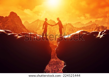 Two men shake hands over precipice between two rocky mountains at sunset. Business, deal, handshake, connection concepts