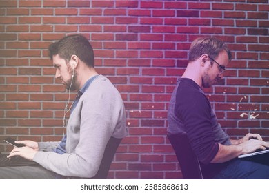 Two men sitting back-to-back, using laptops. Casual attire, focused expressions. Brick wall background. Technology, concentration, work, laptops. Business image.