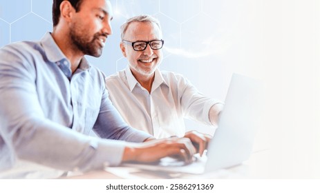 Two men working on laptops, collaborating. One older with glasses, one younger. Business teamwork, technology focus. Bright office setting, teamwork in action. Business image.