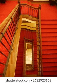 A vertiginous view looking down into a narrow stairwell with ornate wood bannisters and a crimson colored carpet covering  the steps and landings.