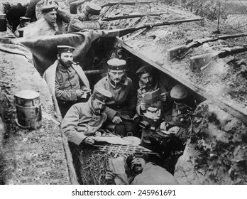 WWI. German soldiers smoking, reading, and writing in their trench. In the background one soldier maintains a battle position. Western Front, ca. 1914-15.