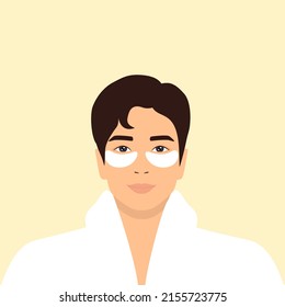Young Asian Man In A White Bathrobe With White Eye Patches On His Face. Daily Skincare Routine
