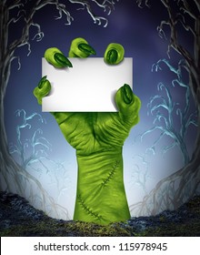 Zombie rising hand holding a blank sign card as a spooky halloween or scary symbol with textured green monster skin in a foggy night tree forest background as a cemetery like creepy place.
