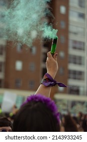 «Bogotá, Colombia»; 03.08.2022: A Woman Wearing A Purple Feminist Scarf Holds In Her Hand A Flare That Releases Green Smoke During The Women's Day March In Colombia»