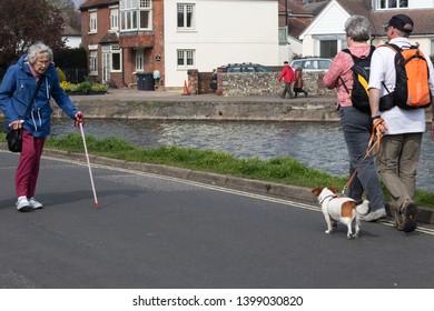 05/03/19 Emsworth, Hampshire, UK An Elderly Walks With Her Walking Stick Past A Middle Aged Couple Walking Their Dog 