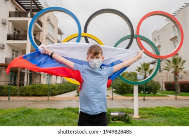 05,04,2021 Russia, Sochi One Person Little Funny Boy In A Medical Mask Stands With The Flag Of Russia Next To The Olympic Rings The Theme Of The Olympic Games