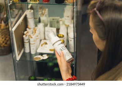 05.28.2022 Warsaw, Poland. Long-haired Caucasian Woman Holding Medical Weed Related Product In One Hand And Reading About Its Benefits. Glass Showcase With Other CBG CBD Products In The Background