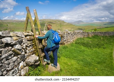 06.05.2022 Settle, North Yorkshire, Uk Woman hill walker wearing green and blue climbs a stile with Pen-y-Ghent in the distance