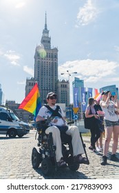 06.25.2022 Warsaw, Poland. Vertical Outdoor Shot Of A Person With Walking Disability Using A Wheelchair In Front Of The Palace Of Culture And Science. LGBTQAI Member Or Ally With Rainbow Flag. High