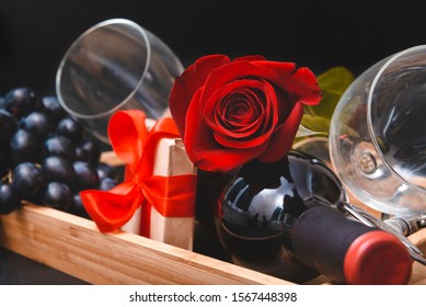 1 Red Rose, Bottle Of Wine, 2 Glasses, Dark Grapes,  Gift Box With Ribbon On Black Background