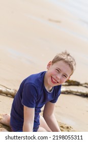 10 11 12 Year Old Boy Sitting On A Beach, Smiling To Camera. Sand Visible Behind Him. Wearing A Tshirt For Swimming. Blonde, Caucasian, Blue Eyes. Summer, Spring, Holiday, Vacation, Sea, Coast, Sand