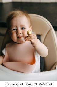 10 months old baby sitting in high chair, with a silicone bib, eating avocado, grimacing. - Shutterstock ID 2150652029