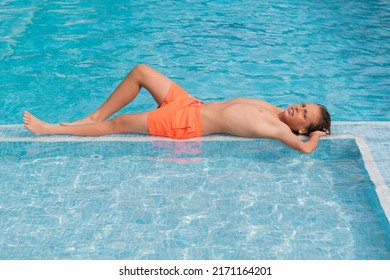 10 Year Old Blond Boy Is Lying In The Pool In The Water, Wearing Orange Swimming Trunks.