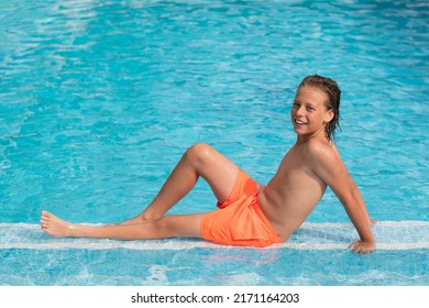 10 Year Old Blond Boy Is Lying In The Pool In The Water, Wearing Orange Swimming Trunks.