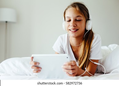 10 years old child using tablet pc. Pre teen girl lying in the bed, listening to music in headphones and browsing social networks. Lazy weekend morning at home.