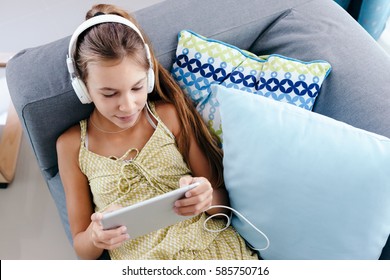 10 years old tween girl relaxing on a couch, listening to music in headphones and playing with tablet pc. Child chilling on the sofa in living room.