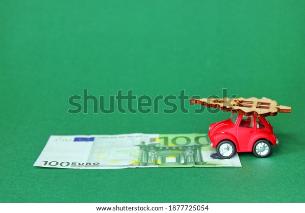 100\
Euros and a red toy car carrying a Christmas tree. A small red toy\
car with a wooden figure of a Christmas tree on the roof. A 100\
Euro bill in front of her on a green\
background.
