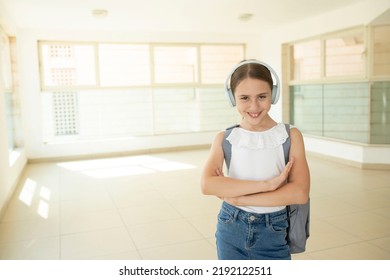 A 10-year-old Schoolgirl Stands In A School Hall With Large Windows. Cute Caucasian Girl With Backpack, Big Headphones. Smiling Face. Concept Of Back To School, Language Courses, Study, Confident Kid.