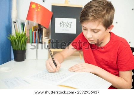 An 11 year old child is learning Chinese. a happy European boy sits at a desk, in front of him is a Chinese flag, on the desk is the hieroglyph “Hello” in Chinese, the boy is doing his homework
