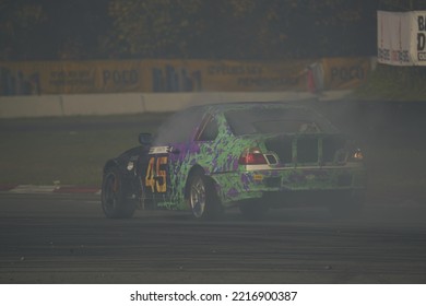 11-05-2022 Riga, Latvia A Car With A Green And Purple Paint Job Driving On A Race Track, A Person On A Race Car In A Track With Smoke Pouring Out The Back.