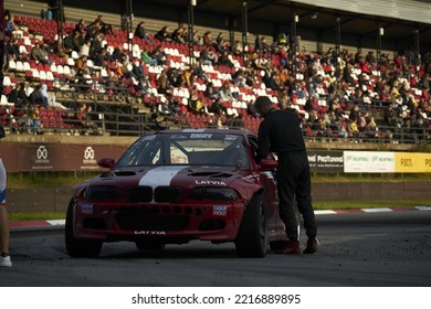 11-05-2022 Riga, Latvia A Man Standing Next To A Racing Car On A Track, A Crowd Looks On As A Race Car Passes By.