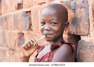 An 11-year-old Ugandan girl smiling, holding a pen against her mouth and leaning against a brick wall looking at the camera
