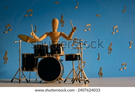 A 12 inch wooden drawing mannequin figure plays on a drum kit against a blue backdrop with gold musical notes and treble and bass clefs