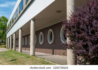 12 Large Round Windows On A Brown Brick Wall In A Straight Row. The Large Windows Have A Grey Concrete Trim Around The Exterior Glass. There's A Walkway Along The Exterior Of The Brick Building.  