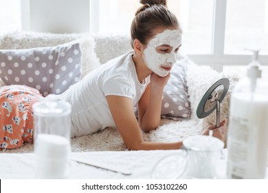 12 years old preteen relaxing on the sofa and making clay facial mask. Teenage girl doing anti blemish skin treatment. Morning skin care routine.