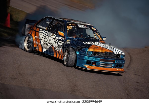 12-05-2022 Riga, Latvia. a blue, orange and black\
race car turning a corner, a race car driving on a track with a\
bunch of smoke coming out of\
it.