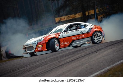 12-05-2022 Riga, Latvia. An Orange And White Race Car Smokes Up A Smoke Trail, An Extreme Sport Car Making Its Way Down The Road.