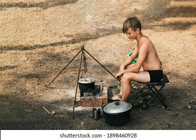 A 12-year-old Boy Is Sitting By A Fire In Nature, Cooking Dinner In A Saucepan Over A Real Fire. Camping Kitchen. A Child Without Clothes On A Hike In Hot Summer. High Quality Photo