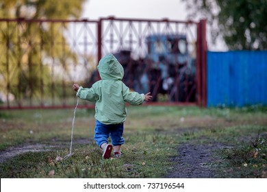 A 1,5 Year Old Kid In A Coat With A Hood  Is Running On The Grass Towards A Tractor Parked Outside The Metal Fence. Kid's Arms Are Raised Up And To The Sides. Back View. Background Is Blurred.