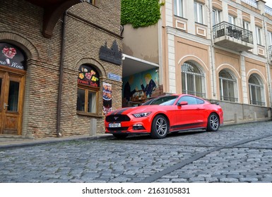 16 May 2022 - Georgia, Tbilisi.
Modern Muscle Car Ford Mustang. Car Is Orange With Black Stripes And Parked Near Old Houses On The Grey Cobblestone Road.