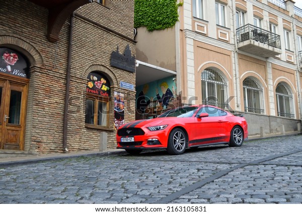16 May 2022 - Georgia, Tbilisi.\
Modern\
muscle car Ford Mustang. Car is orange with black stripes and\
parked near old houses on the grey cobblestone\
road.