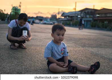 18th March 2018 Chanthaburi Province Thailand : Cute Small Child Sitting On A Ground With Mother Taking Photo With Smartphone From Behind During Evening Sunset