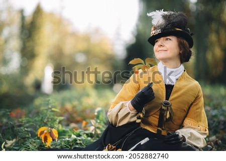 A 19th century woman sits on the grass in a park holding an autumn leaf in her hand