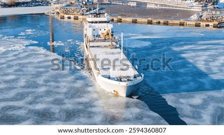 1A ice class general cargo ship manoeuvring while leaving industrial port. Sea surface covered in thin ice. Aerial front view.